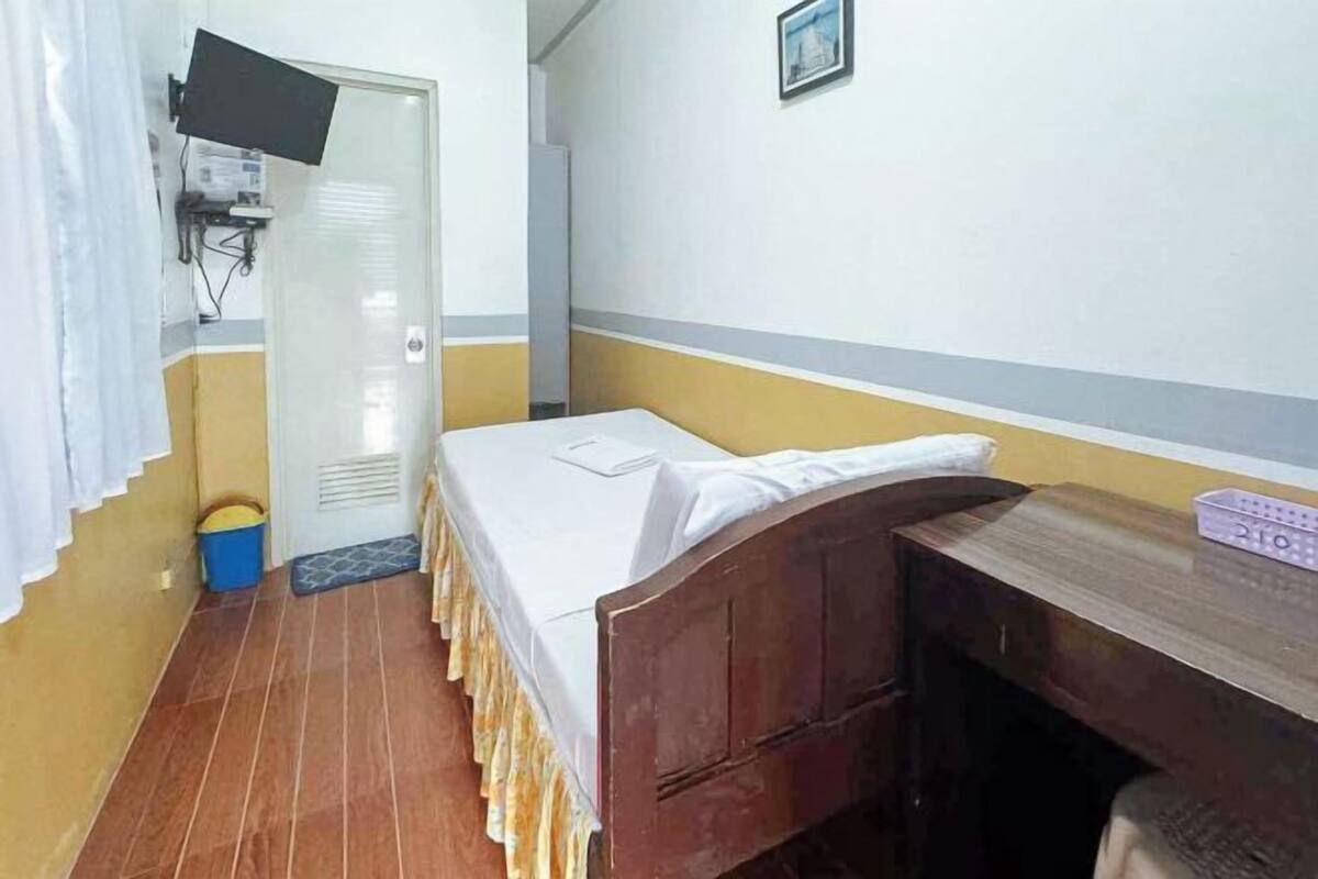 A small, single-bed room with a wooden headboard, a desk, a television mounted on the wall, and a door with a window.