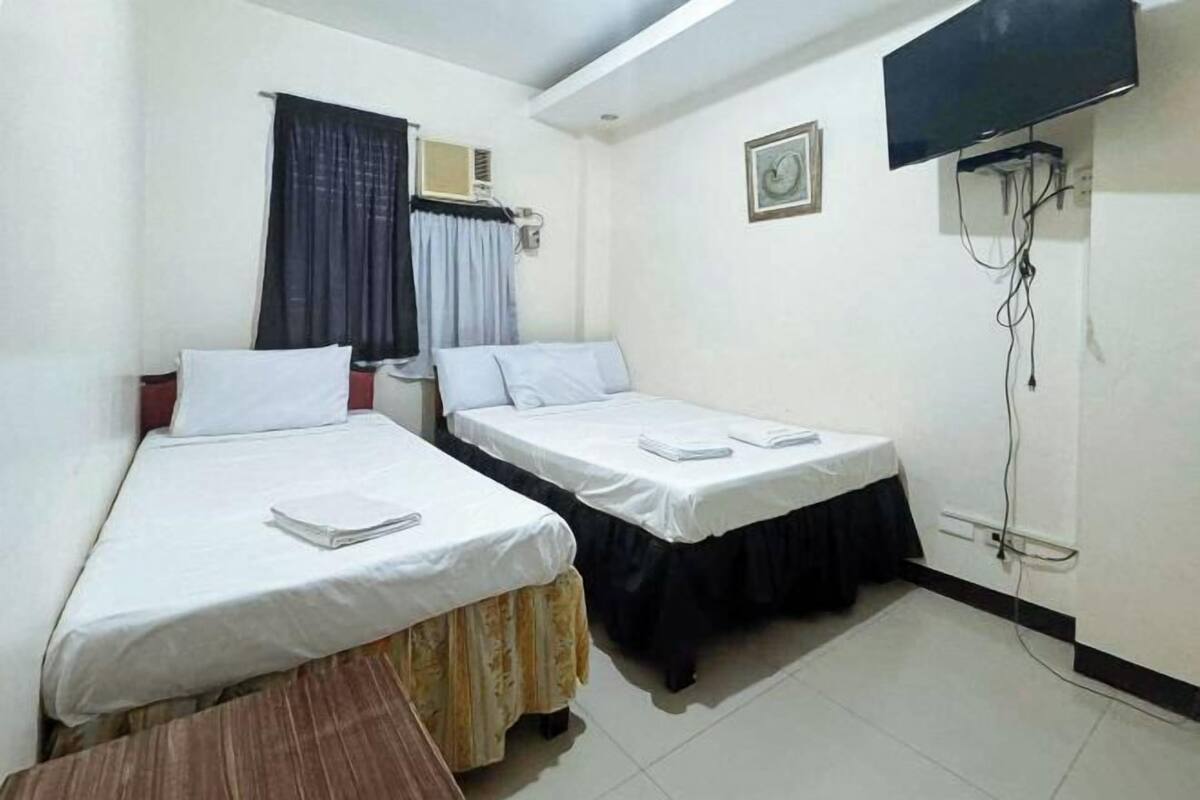 A hotel room with two single beds, a small table, a TV mounted on the wall, and an air conditioning unit.