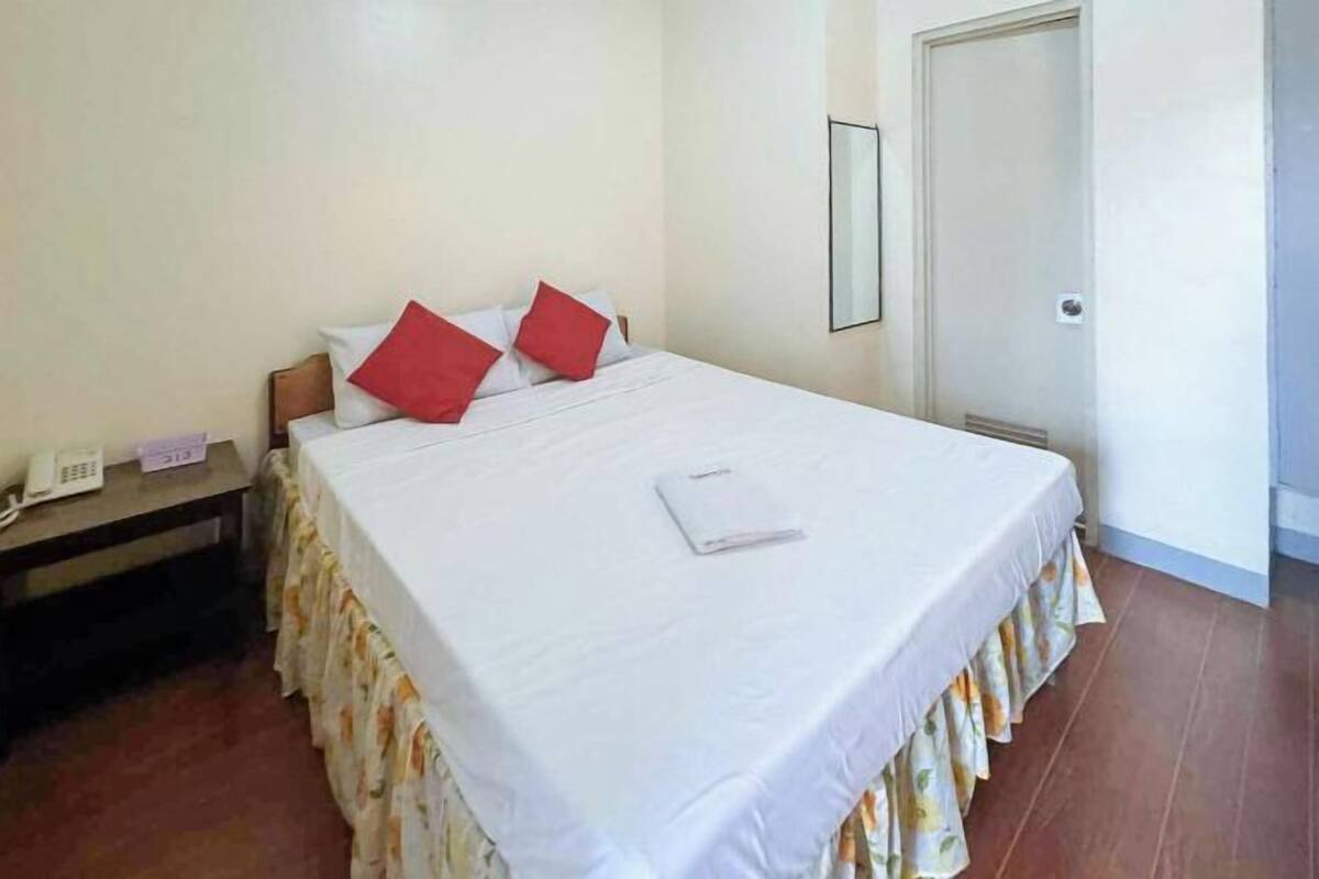 A neatly made bed with white bedding and red pillows, a bedside table with a phone and a small item, and a door in the background.