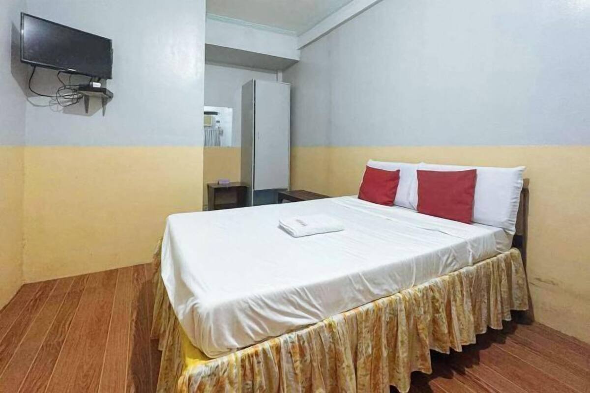 A single bed with a white bedspread and a floral skirt, a flat-screen TV mounted on the wall, and a refrigerator in the background.