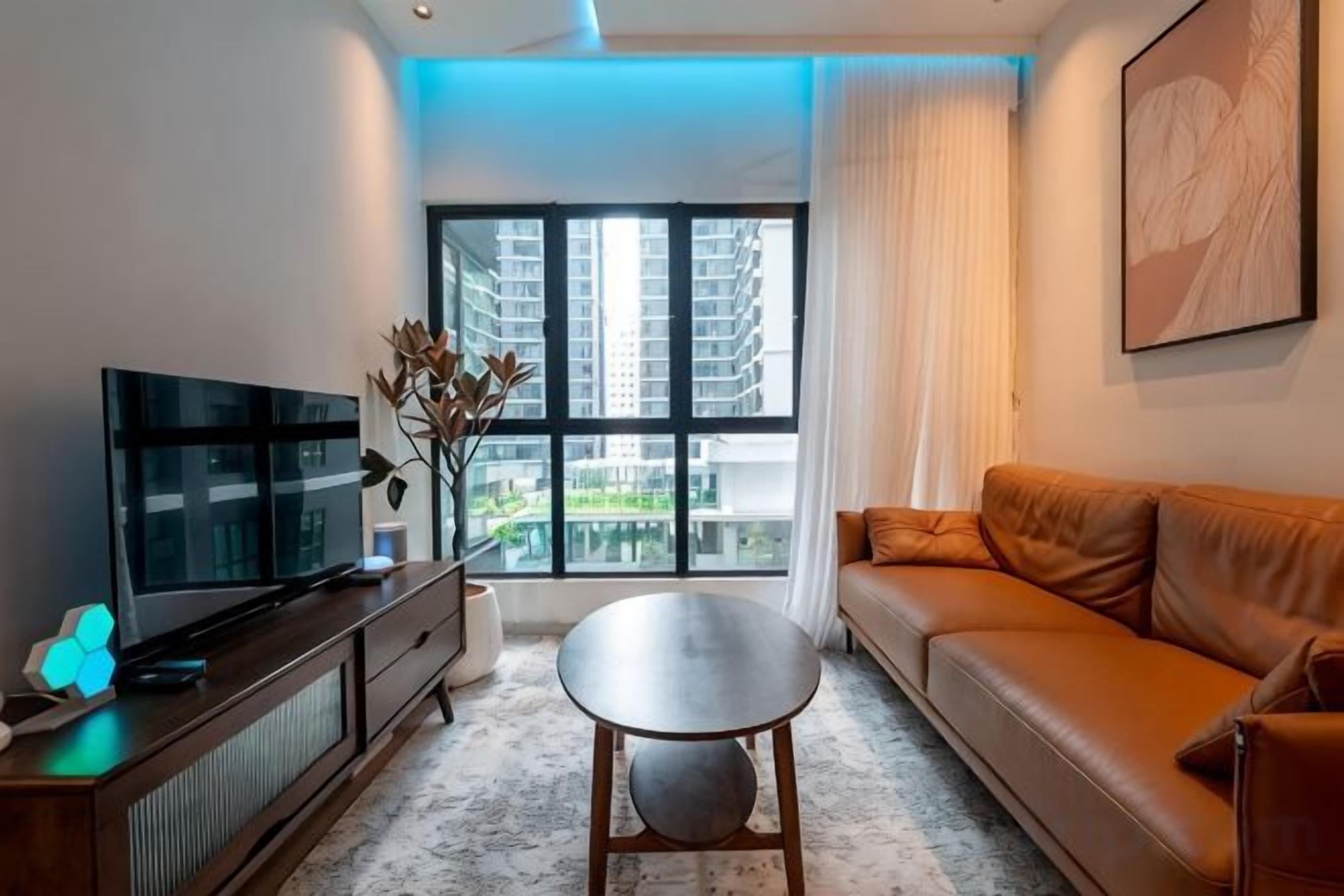 A modern living room with a flat-screen TV, a leather sofa, a round coffee table, and a large window offering a city view.