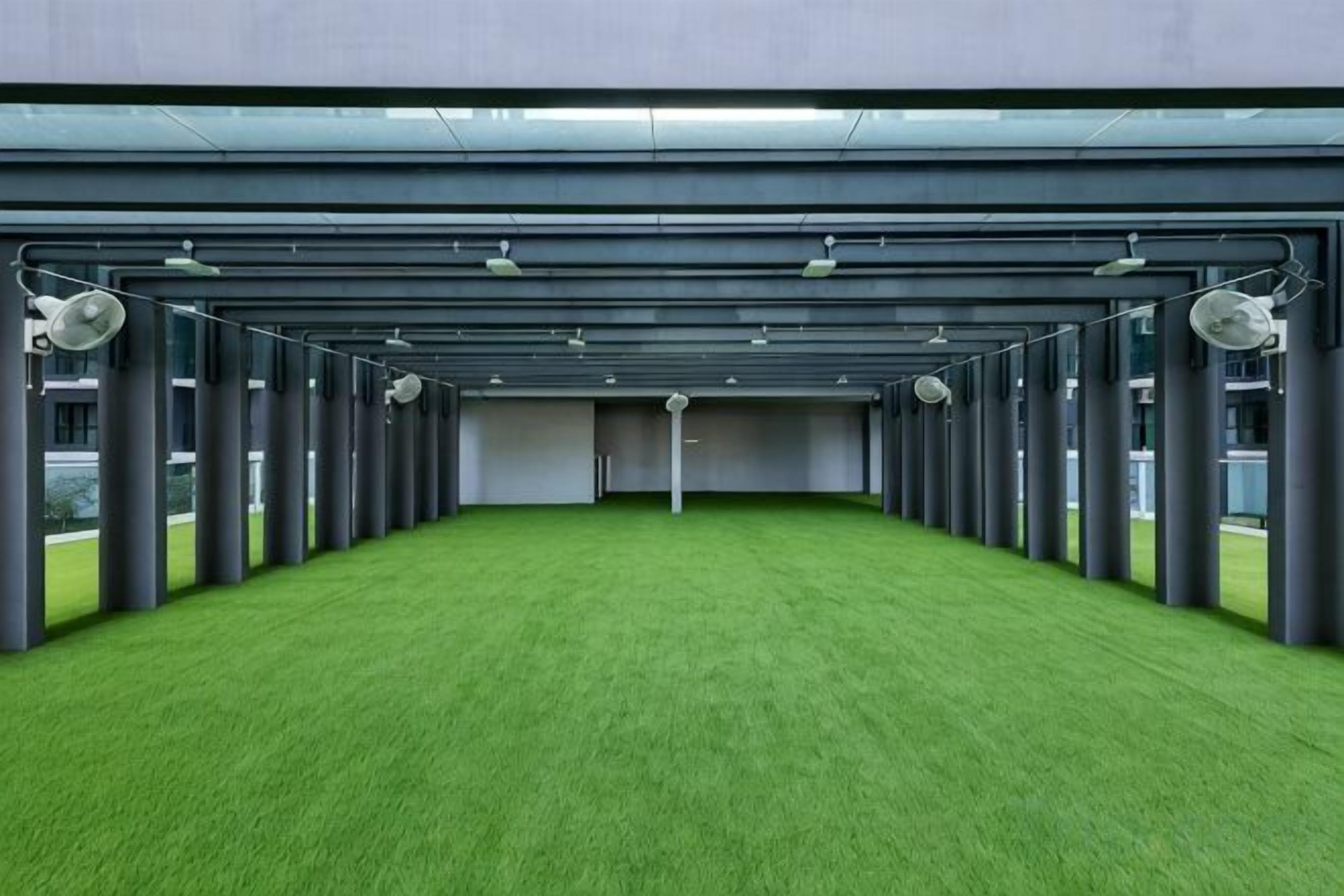 Un amplio espacio interior con suelo de césped artificial verde, múltiples luces en el techo y una fila de pilares.