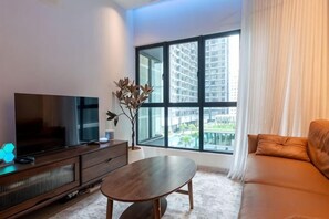 A modern living room with a flat-screen TV, a wooden coffee table, a sofa, and a large window offering a view of high-rise buildings. - MAXIM BY FEEL HOME (Kuala Lumpur)
