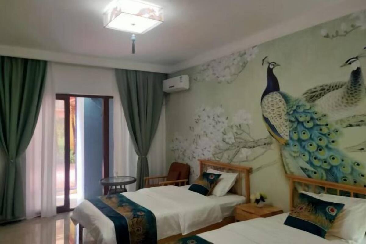 A hotel room with two beds, a peacock mural, a window with curtains, and an air conditioning unit.