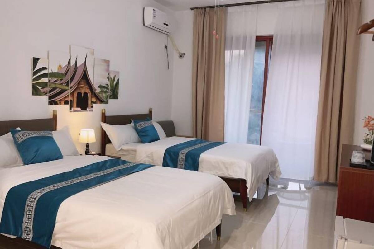 A hotel room with two beds, a desk, a chair, a lamp, a wall-mounted air conditioner, and a window with curtains.
