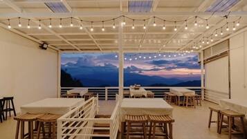 Un área de comedor al aire libre con mesas y sillas, luces colgantes y una vista panorámica.