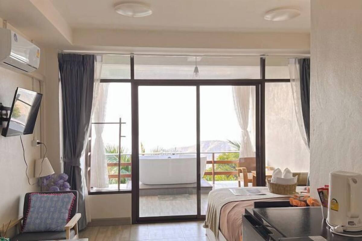 A hotel room with a bed, a desk, a chair, a TV, and a balcony with a view of greenery.
