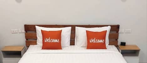Una cama bien hecha con ropa de cama blanca y dos almohadas naranjas con el texto 'welcome', flanqueada por dos mesitas de noche con lámparas.