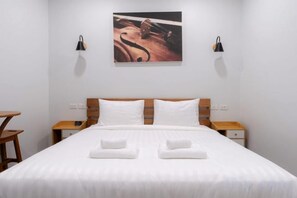 Un lit bien fait, avec du linge de lit blanc et deux serviettes, une tête de lit en bois, des tables de chevet avec des lampes et une œuvre d’art accrochée au mur.
