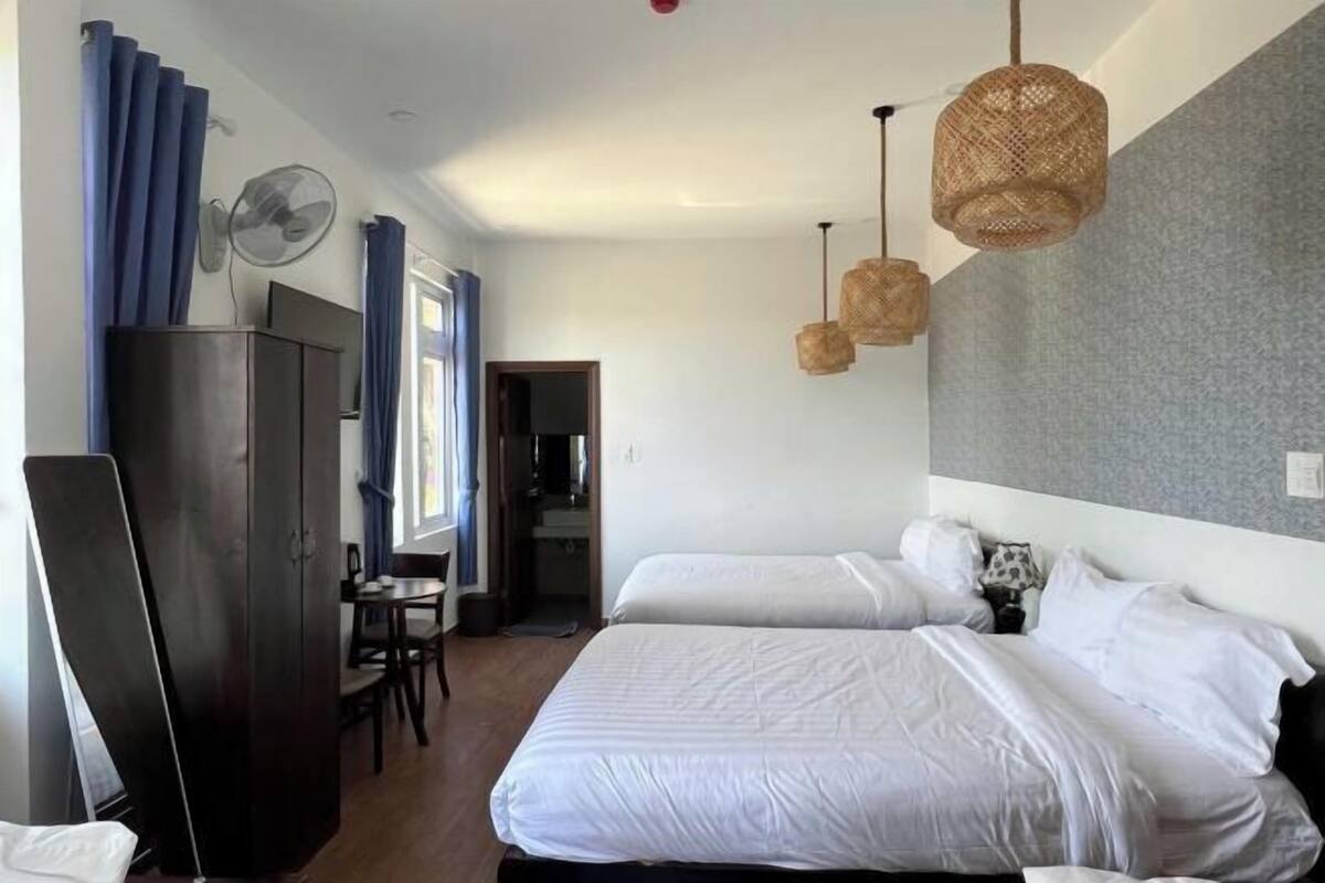 A hotel room with two beds, a desk, a chair, a wardrobe, and a fan.