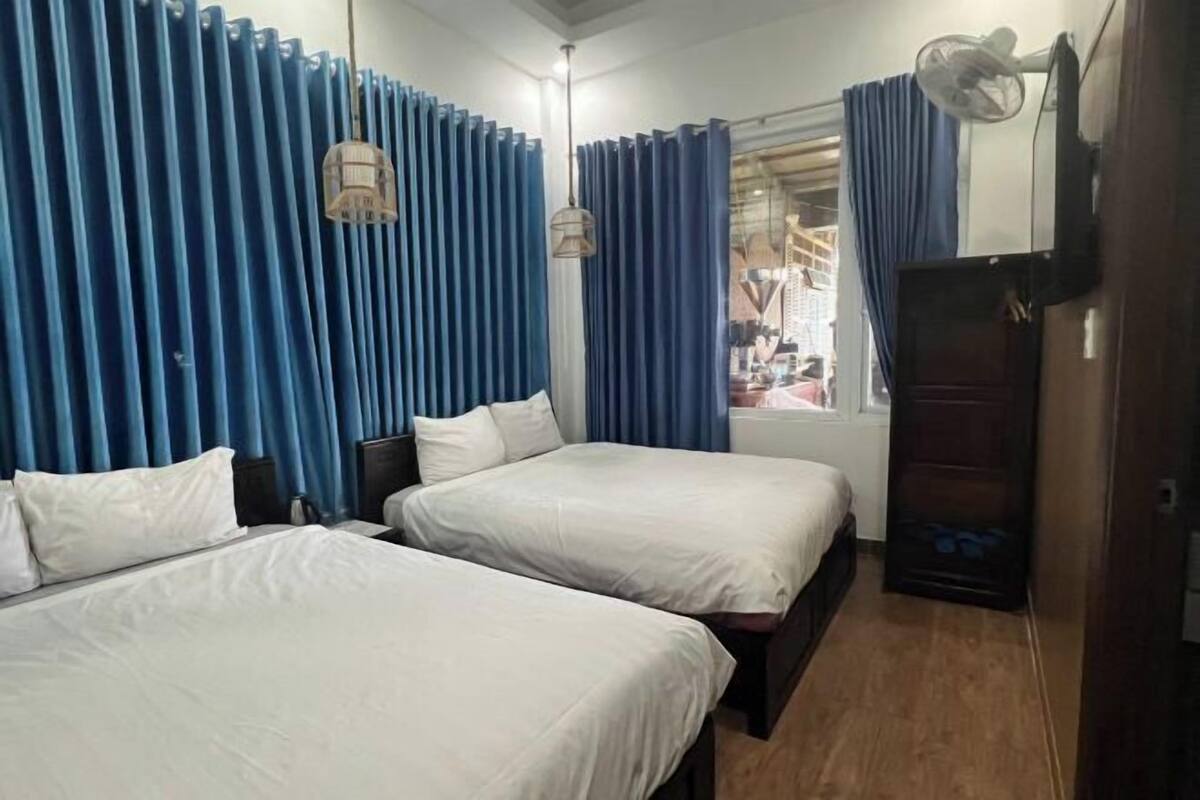 A hotel room with two beds, a wardrobe, a desk, a chair, a window with curtains, and a fan.