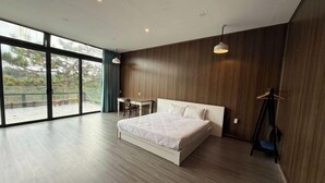 A modern bedroom with a large bed, a desk, and a view through large windows. - Michelia Valley Hotel (Dalat)