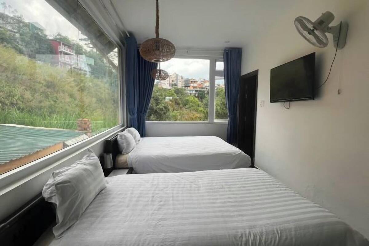 A room with two beds, a TV, a wall-mounted fan, and a window with curtains.