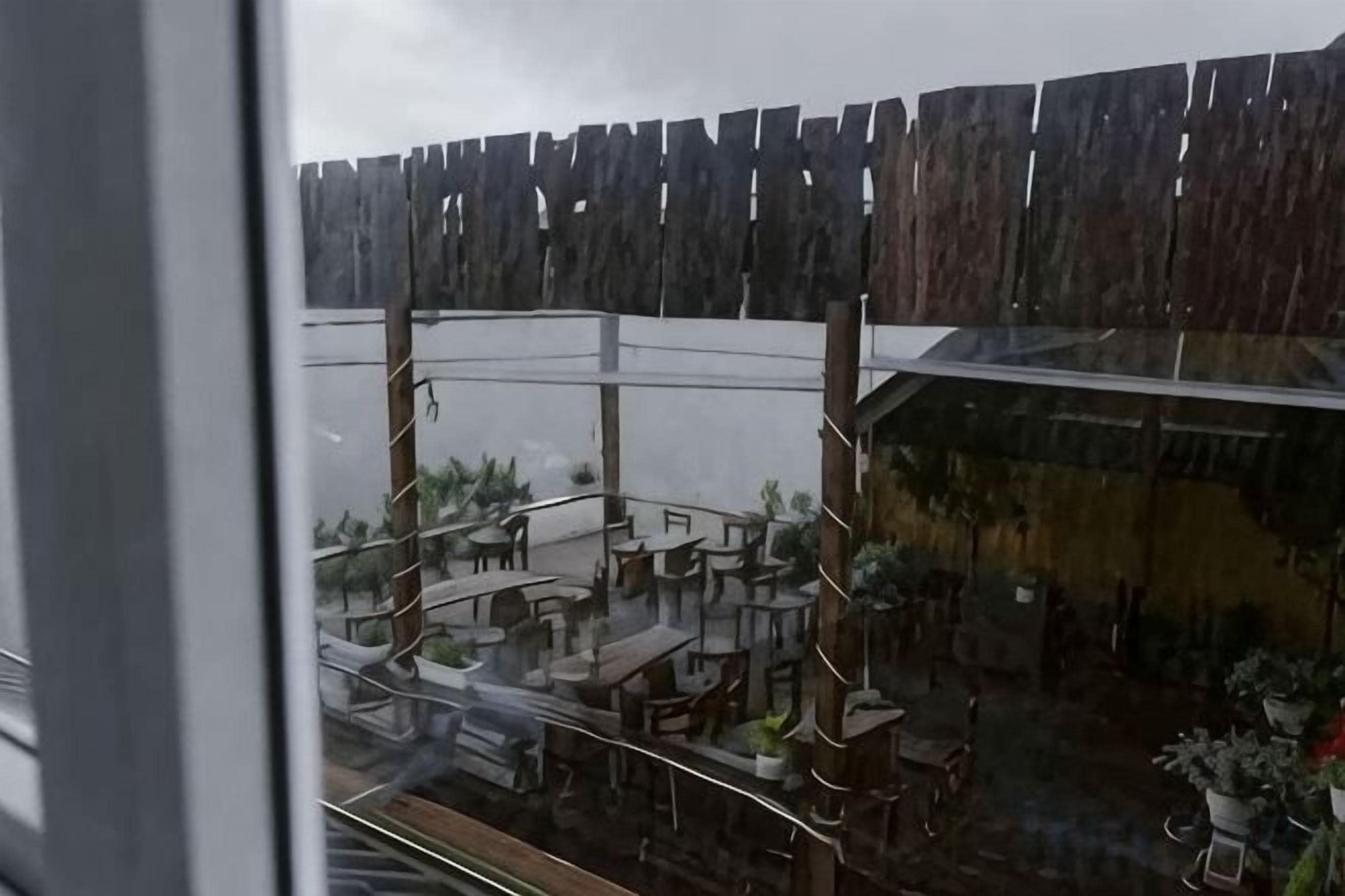 A view of a covered outdoor seating area with tables and chairs, a wooden fence, and potted plants.