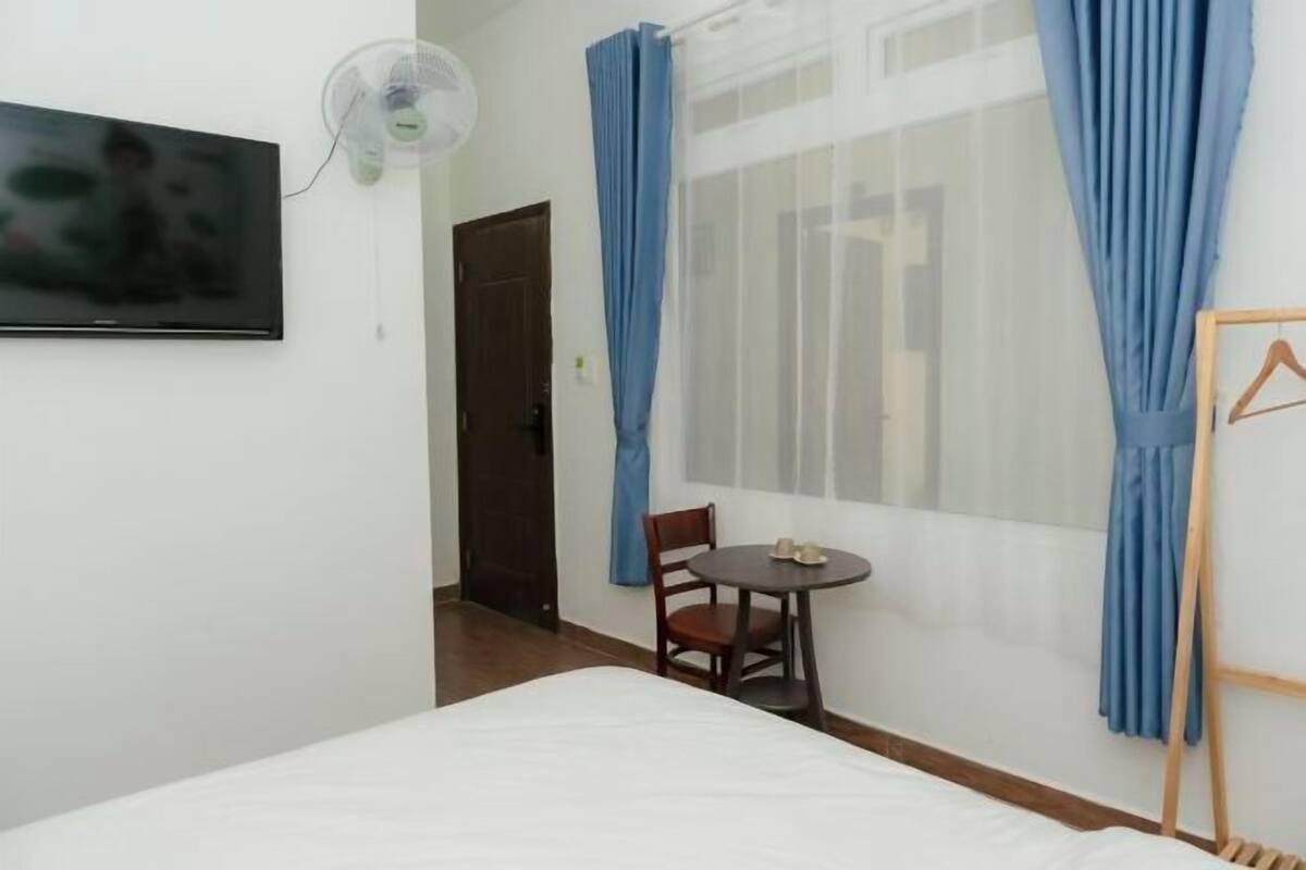 A hotel room with a bed, a TV, a fan, a table with a chair, and a window with curtains.