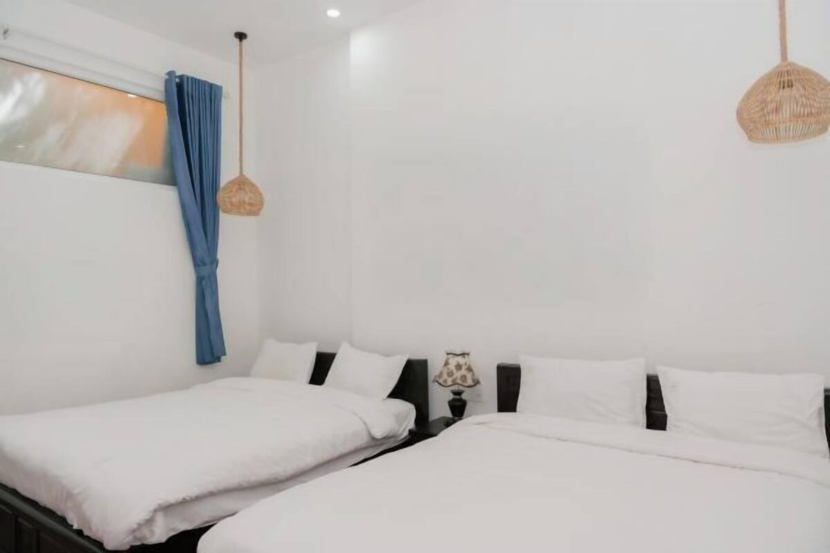 A room with two beds, white bedding, a window with blue curtains, and two hanging wicker lamps.