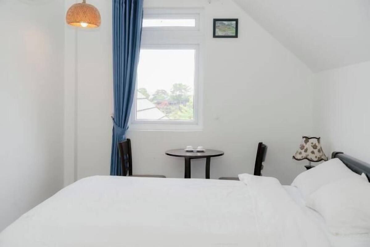 A bedroom with a bed, a small table, a chair, a lamp, a window with curtains, and a hanging light fixture.
