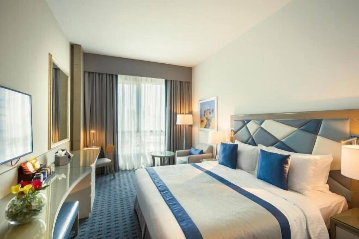 A hotel room with a bed, a desk, a chair, a lamp, and a window with curtains.