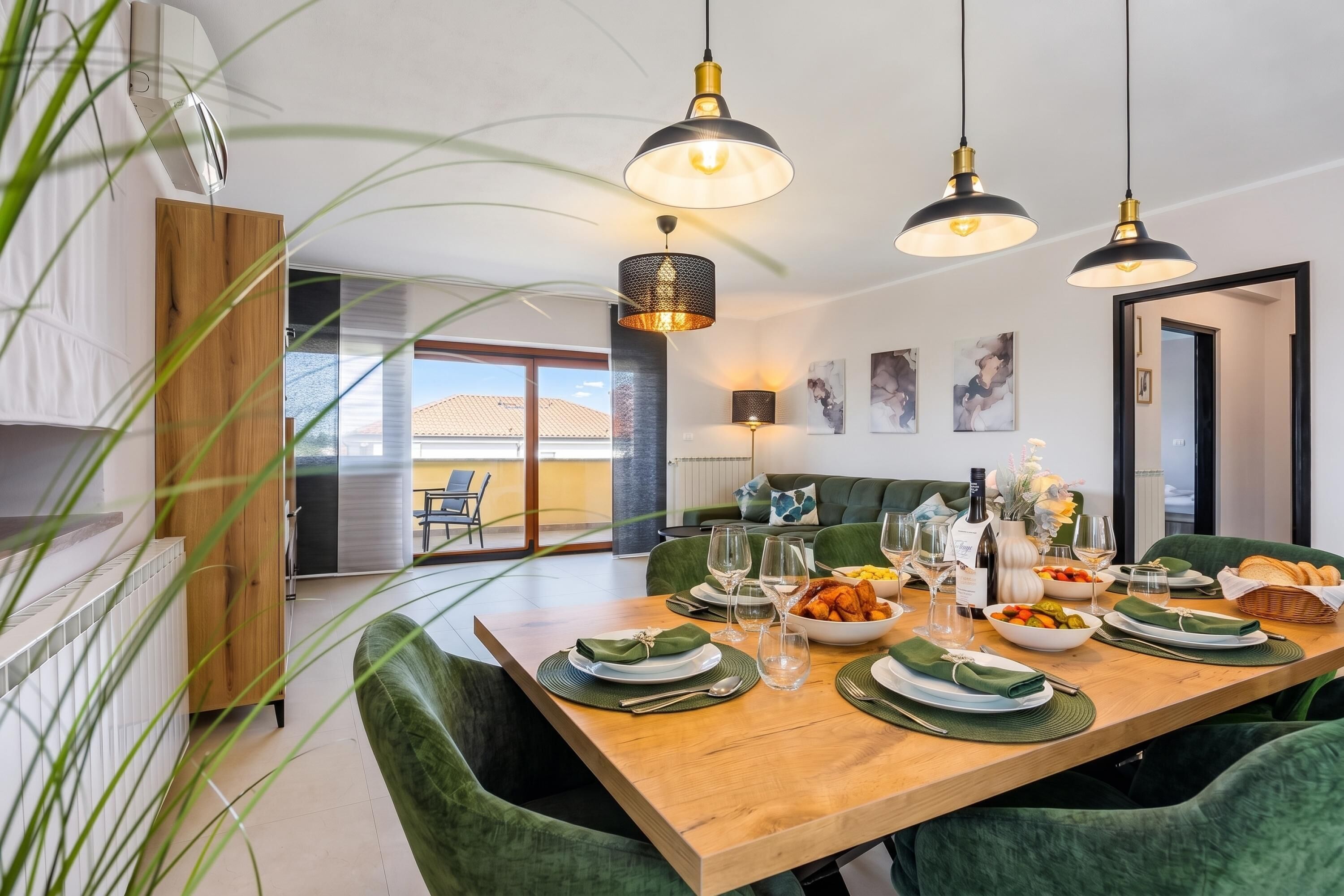 A modern dining area with a wooden table set for a meal, green upholstered chairs, and pendant lighting.