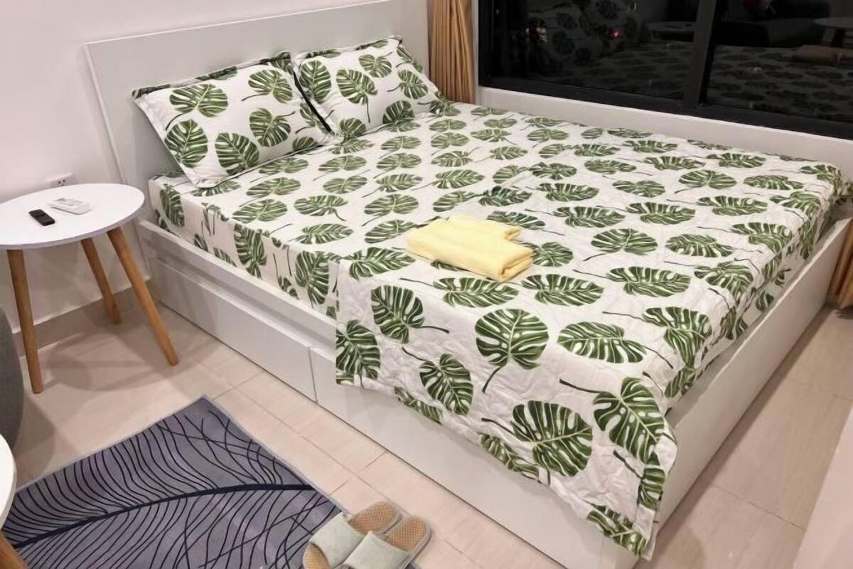 Una cama con una colcha estampada con motivos de hojas, una mesita de noche, una alfombra pequeña y pantuflas.