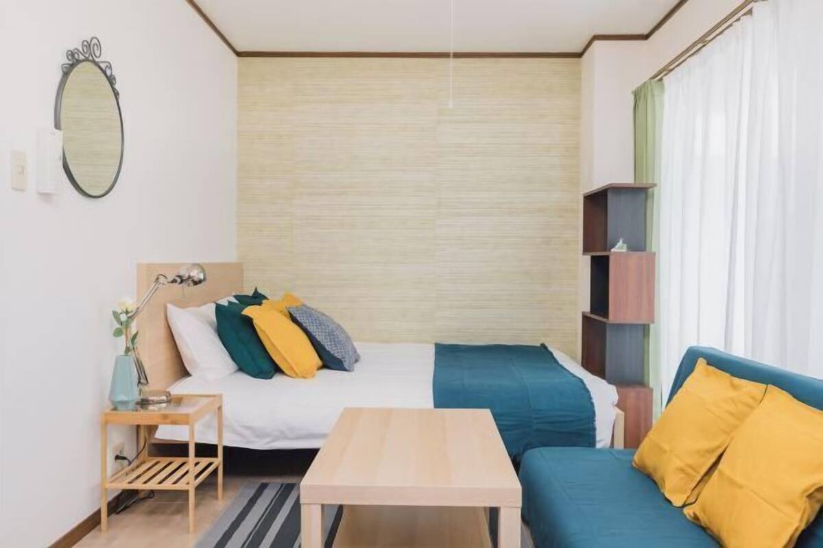 A compact room with a bed, a sofa, a small table, a mirror, and a shelf unit.
