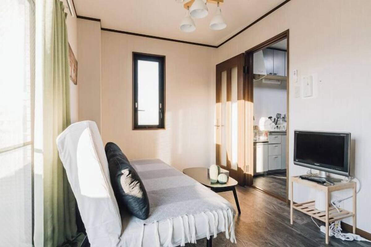 A hotel room with a bed, a TV on a stand, a small table with a lamp, and a view of the kitchen through an open door.