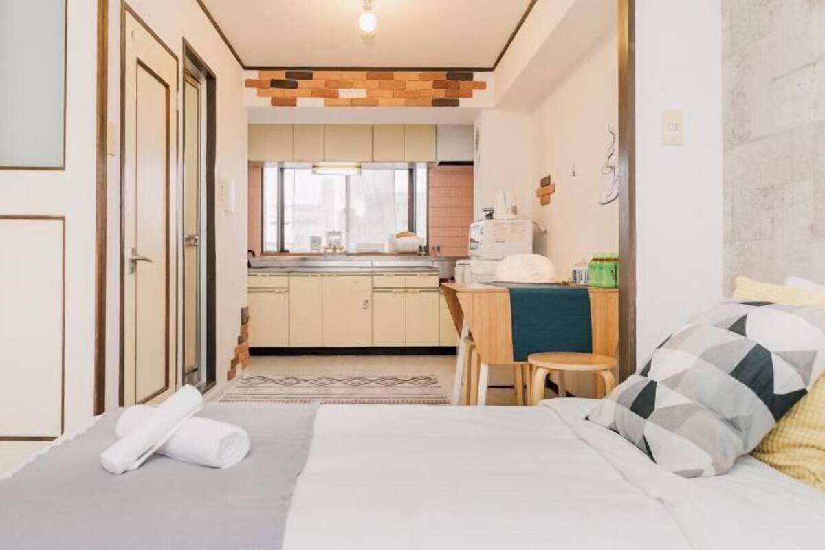 A compact hotel room with a bed, a small desk, a chair, a kitchenette, and a bathroom.