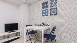 A small dining area with a white table and two blue chairs, a television on a white stand, and a wall with patterned tiles. - Nippombashi Back Kuromon AFP Apartment (Osaka)