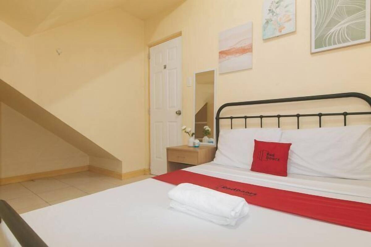 A hotel room with a bed, a red throw, a white pillow, a red pillow with text, a nightstand, a mirror, and a staircase.