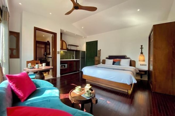 A hotel room with a bed, a sofa, a small table with cups, a ceiling fan, and a view of the kitchen.