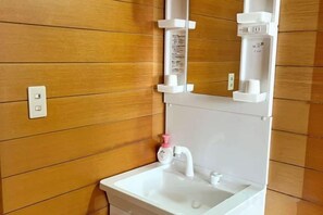 A bathroom with a wooden wall, a white sink, and a mirror with a soap dispenser. - FujiAlpes (Fujikawaguchiko)