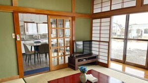 Une pièce de style japonais traditionnel, avec un sol recouvert de tatamis, une petite table en bois, un téléviseur et des portes coulissantes donnant sur une cuisine.