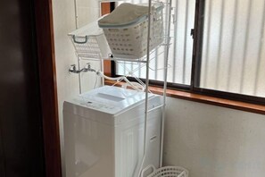 A washing machine with a basket on top, a window with blinds, and a white wall. - FujiAlpes (Fujikawaguchiko)