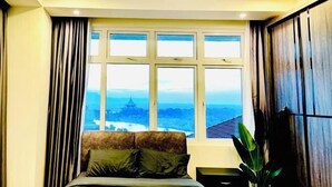 A bedroom with a large window, a bed, a bedside table, and a plant. - Kuching City Center Riverine Apartment With Balcony & City Views (Kuching)