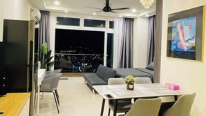 A modern living room with a sofa, dining table, and chairs. There is a TV, a ceiling fan, and a chandelier. The room has large windows with curtains. - Kuching City Center Riverine Apartment With Balcony & City Views (Kuching)