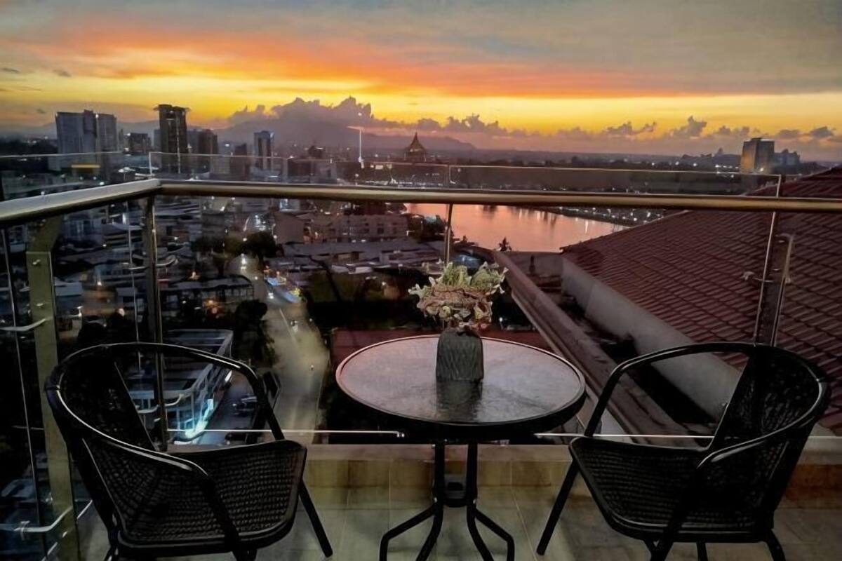 A balcony with two chairs and a table, overlooking a cityscape and a river.