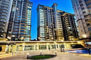 Un hotel moderno con varios edificios altos al fondo, una entrada bien iluminada y una zona ajardinada en primer plano.