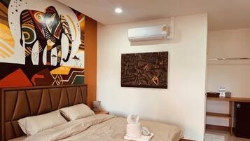 A hotel room with a bed, a wall-mounted air conditioner, a painting, and a kitchenette area.