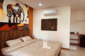 A hotel room with a bed, a wall-mounted air conditioner, a painting, and a kitchenette area.