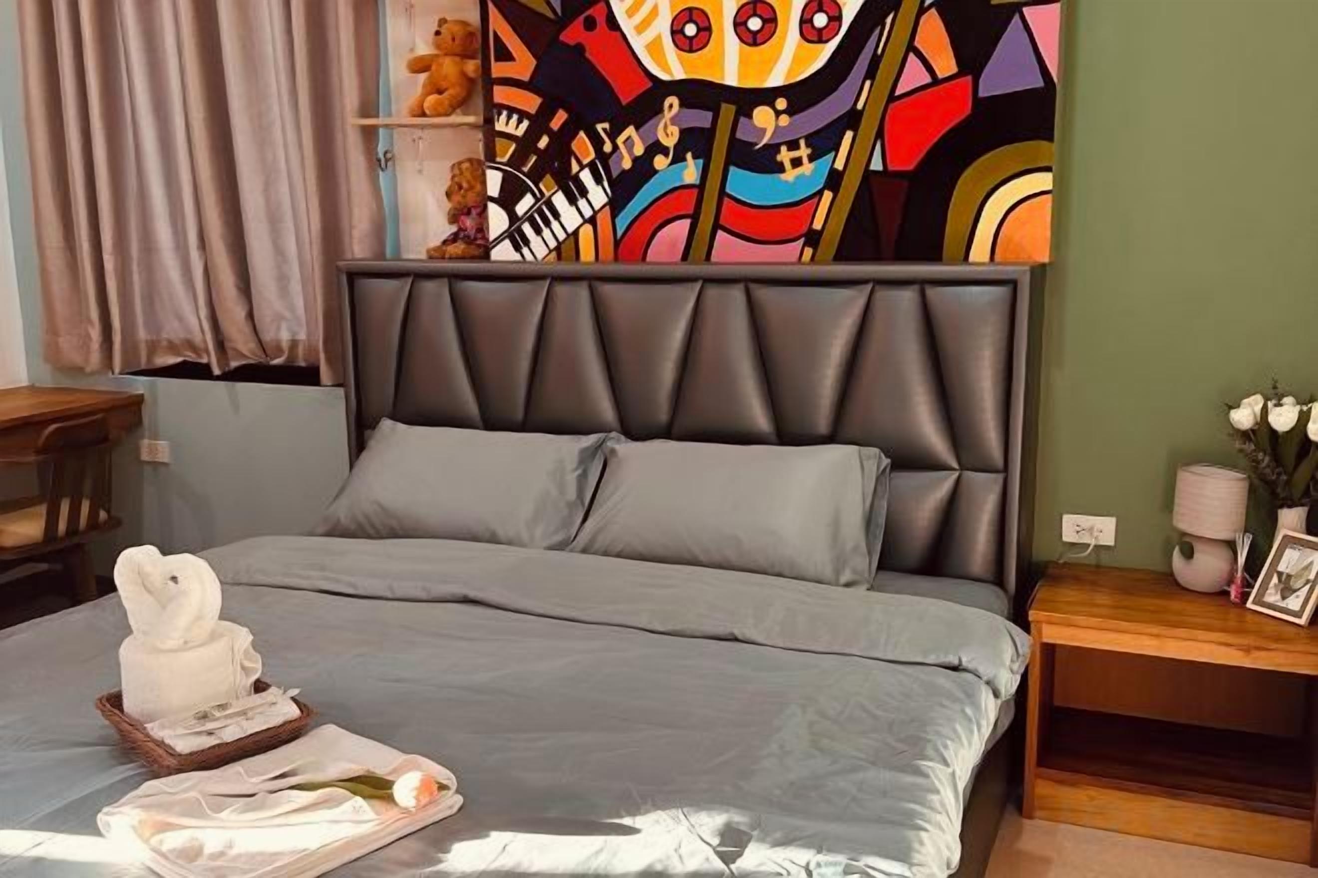 Un lit avec une tête de lit grise et des oreillers, une table de chevet avec un vase et un cadre photo, et une peinture abstraite aux couleurs vives accrochée au mur.