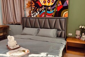Una cama con cabecera gris y almohadas, una mesita de noche con un florero y un marco de fotos, y un cuadro abstracto de colores en la pared.