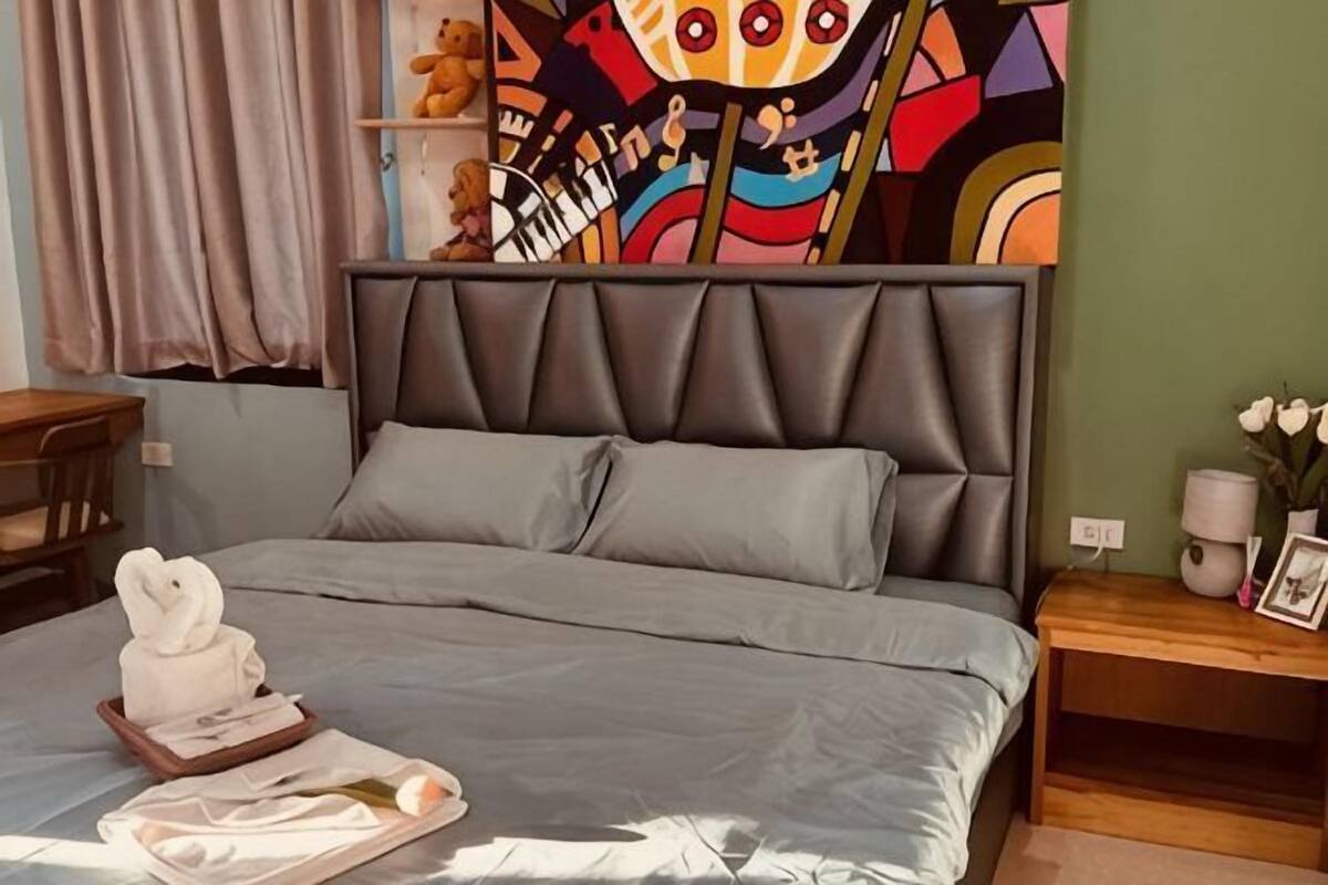 Een slaapkamer met een grijs bed, een hoofdbord, een nachtkastje en een kleurrijke wandtapijt.
