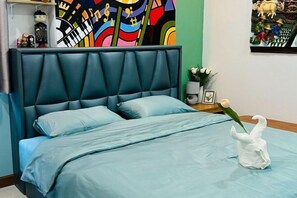 Una cama con cabecera azul, una colcha azul y un jarrón blanco con forma de cisne que contiene una sola rosa.