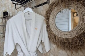 A white robe hanging on a rack, a woven wall hanging, and a round mirror with a wooden frame. - Mahkota Sangkuriang Bandung (Cisarua)