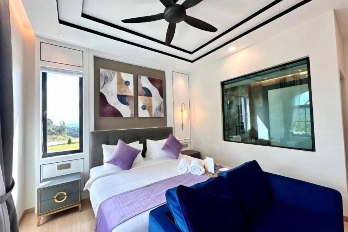 A modern hotel room with a large bed, a ceiling fan, artwork on the wall, and a view through a window.