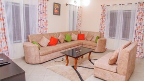 A living room with a beige sectional sofa, a glass coffee table, and a small armchair. There is a remote control on a table, and curtains with a geometric pattern are on the windows. - Tremendous 4-bedroom bungalow in Kilifi County (Kilifi County)