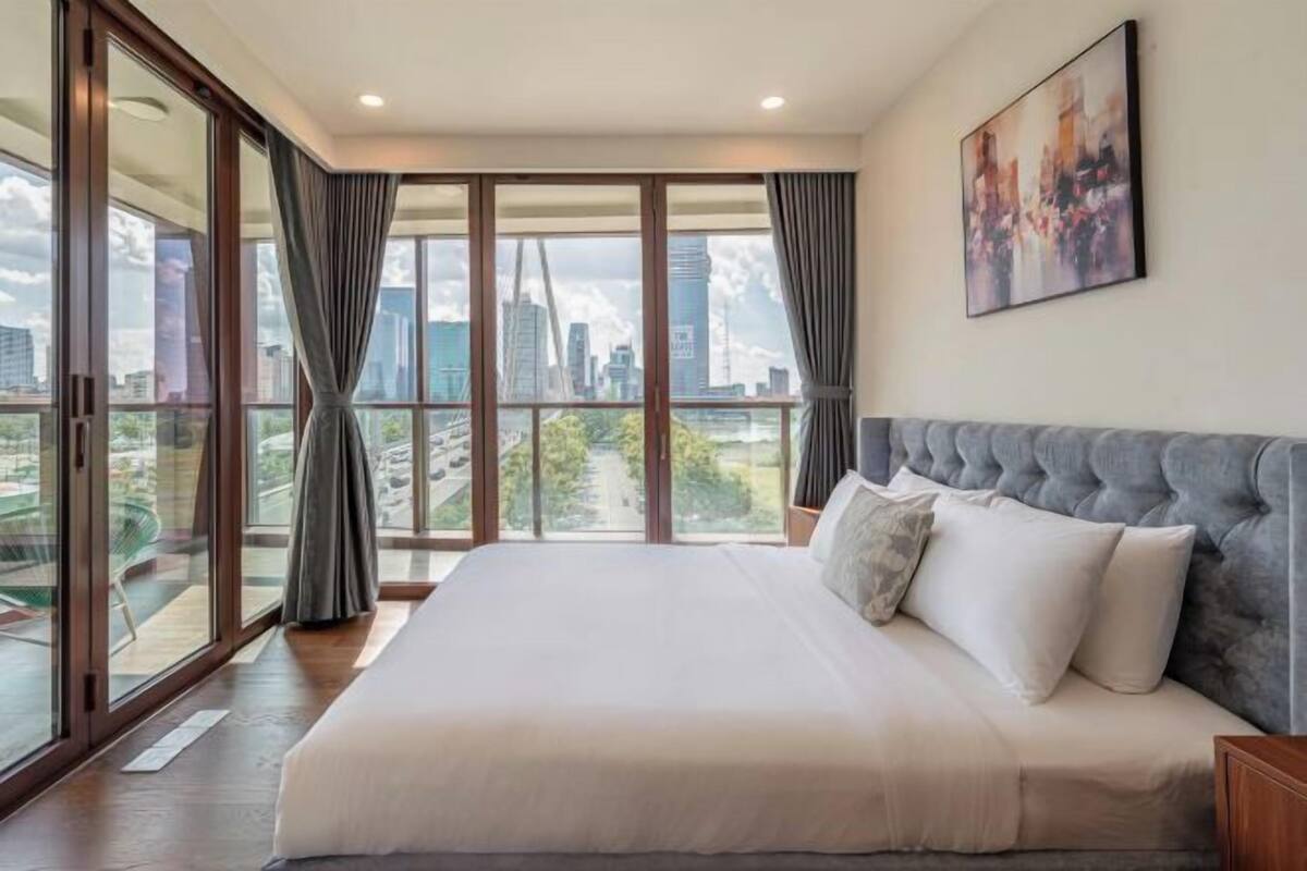 A bedroom with a large bed, a city view through floor-to-ceiling windows, and a painting on the wall.