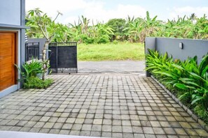 Un camino pavimentado conduce a una entrada con portón, rodeado de exuberante vegetación y una fachada de edificio moderno.