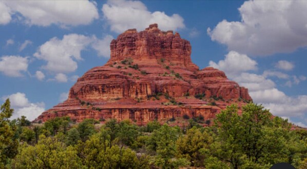 Una gran formación rocosa roja de cima plana, rodeada de árboles verdes.