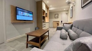 A modern living room with a flat-screen TV, a grey sofa, a wooden coffee table, and a kitchen area in the background. - LV Modern Suite Langkawi by Zervin (Langkawi)